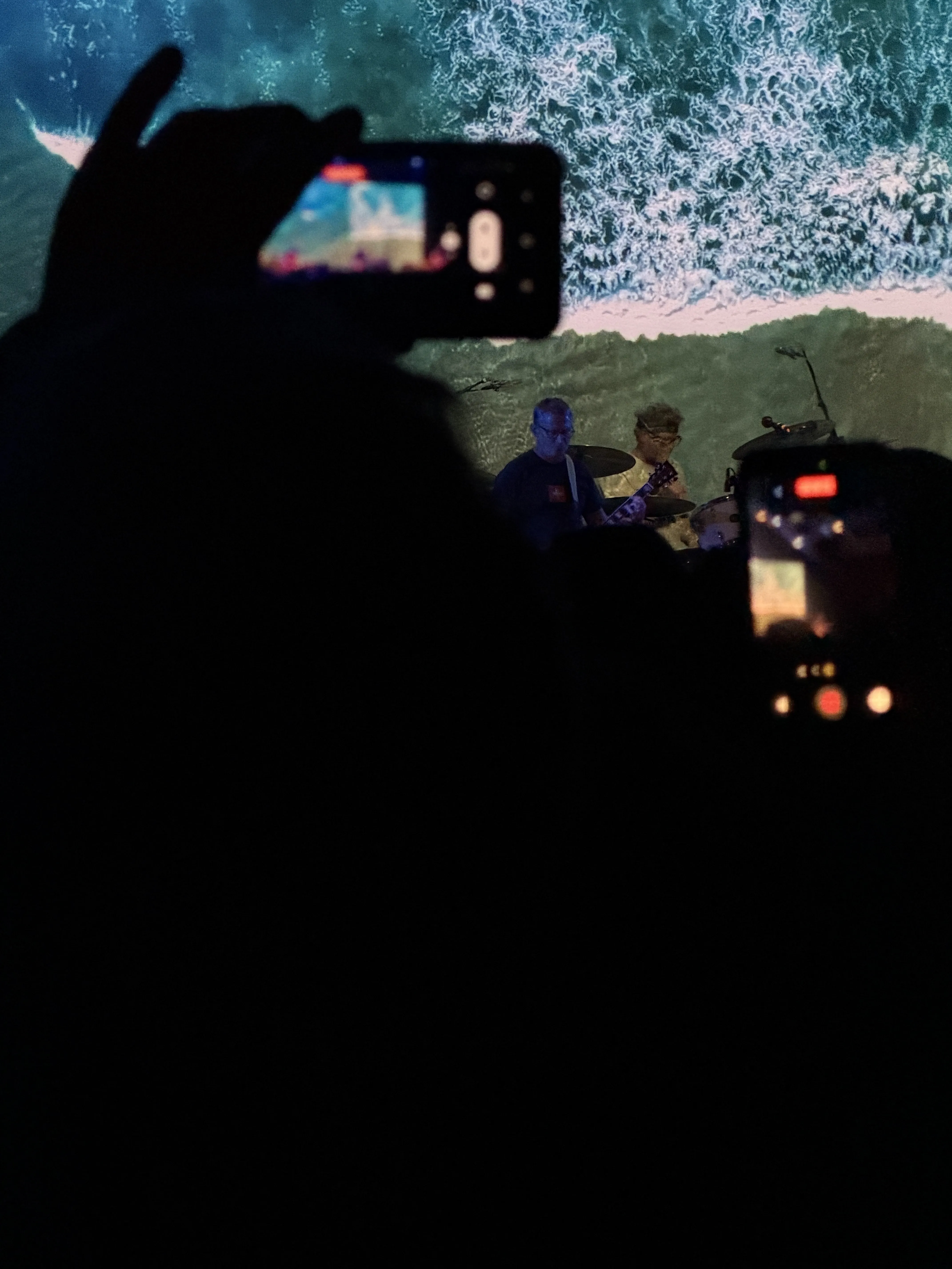 Hum performing during Slide Away fest in Los Angeles — silhouetted phones held up in the foreground, an ocean wave projected behind the band