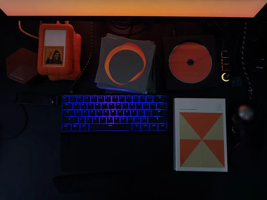 Desk with LaCie rugged drive, Alva Noto CDs, the Alva Noto + Ryuichi Sakamoto V.I.R.U.S. 5 CD box set reissue, and a blue-lit mechanical keyboard, monitor glowing orange in the background