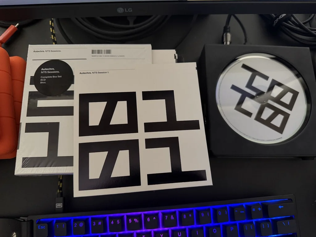 Autechre NTS Sessions complete box set and NTS Session 1 CD on a desk, with a blue-lit mechanical keyboard in the foreground and an LG monitor in the background