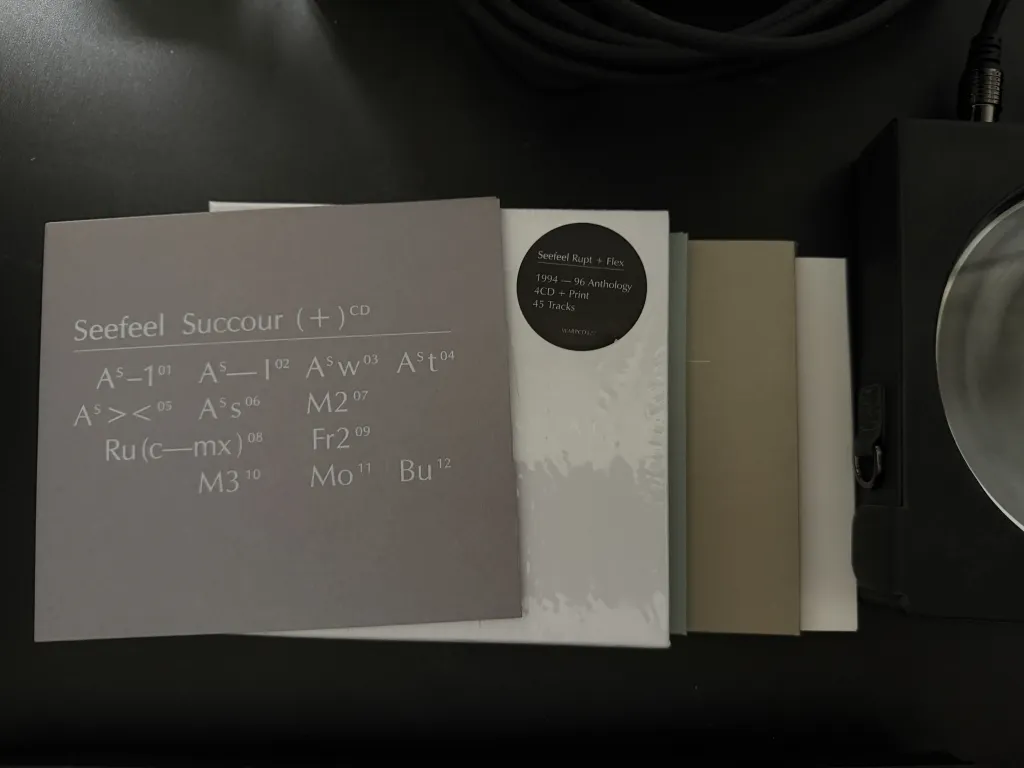 Seefeel — Succour (+) CD, its inner sleeve printed with the album's abbreviated tracklist, beside the shrink-wrapped Seefeel Rupt + Flex 1994–96 Anthology 4CD box set on a black desk, with a turntable platter at the right edge