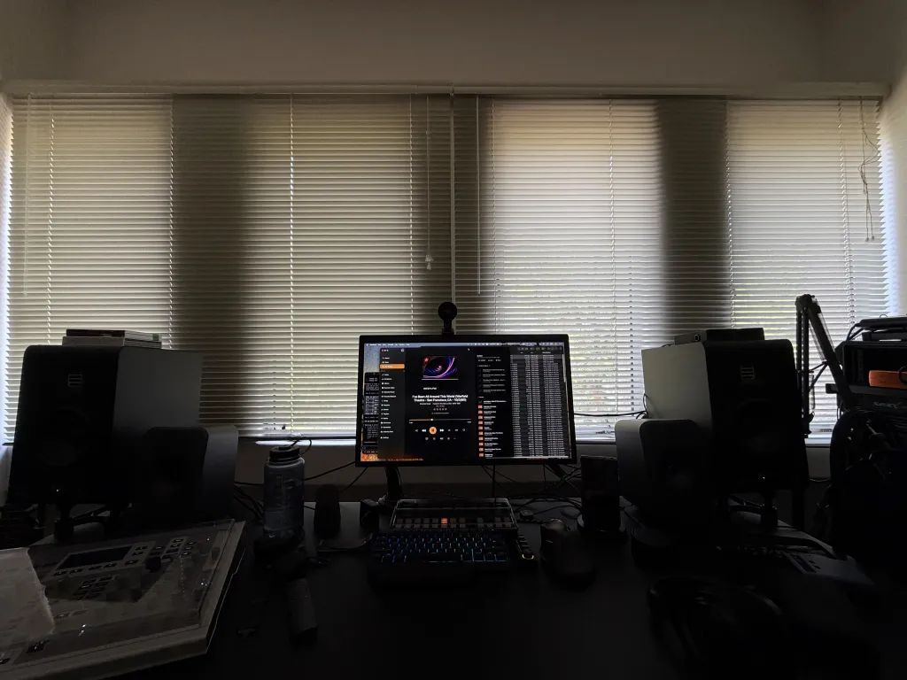 Dim studio at dusk, venetian blinds glowing behind a monitor showing Swinsian playing Grateful Dead's 'I've Been All Around This World' from Hunter's Trix Bonus Disc 1975-1981, flanked by studio monitors and a mixing console in the foreground
